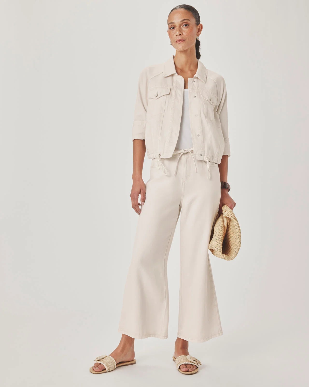 Woman wearing a beige outfit with wide-leg pants and a jacket, holding a hat against a white background