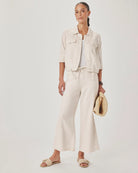 Woman wearing a beige outfit with wide-leg pants and a jacket, holding a hat against a white background