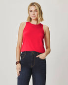 Woman wearing a red sleeveless top and dark jeans on a plain background