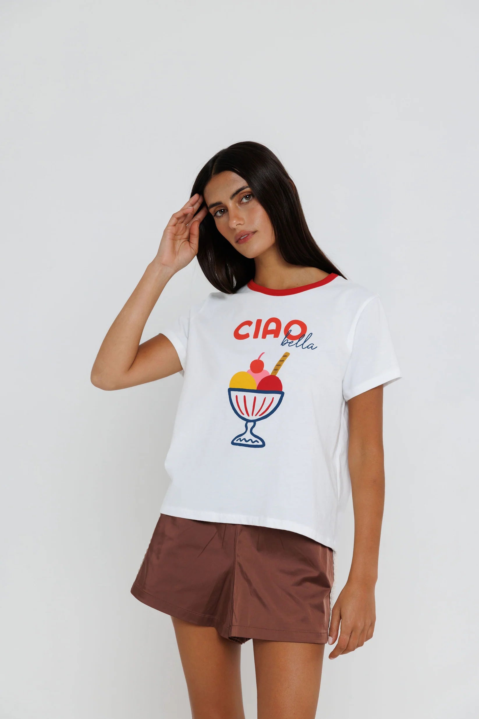 A woman modeling a white short-sleeve t-shirt with a red collar trim and a colorful 'CIAO bella' graphic on the front, paired with brown shorts, against a light gray studio background.