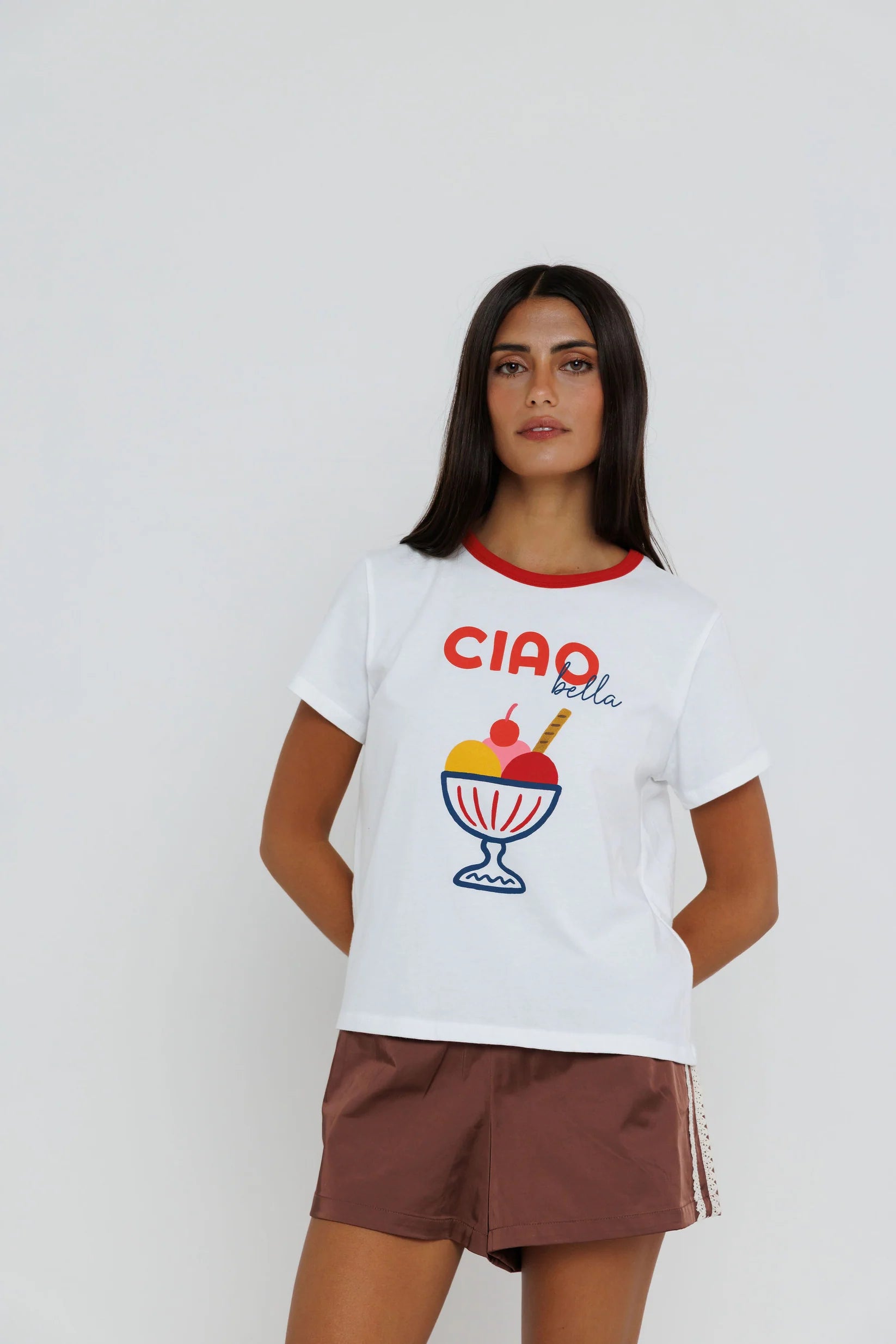 A woman modeling a white short-sleeve t-shirt with a red crew neck trim, featuring a colorful graphic of a sundae and the text 'CIAO bella' on the front, paired with brown shorts against a light gray background.