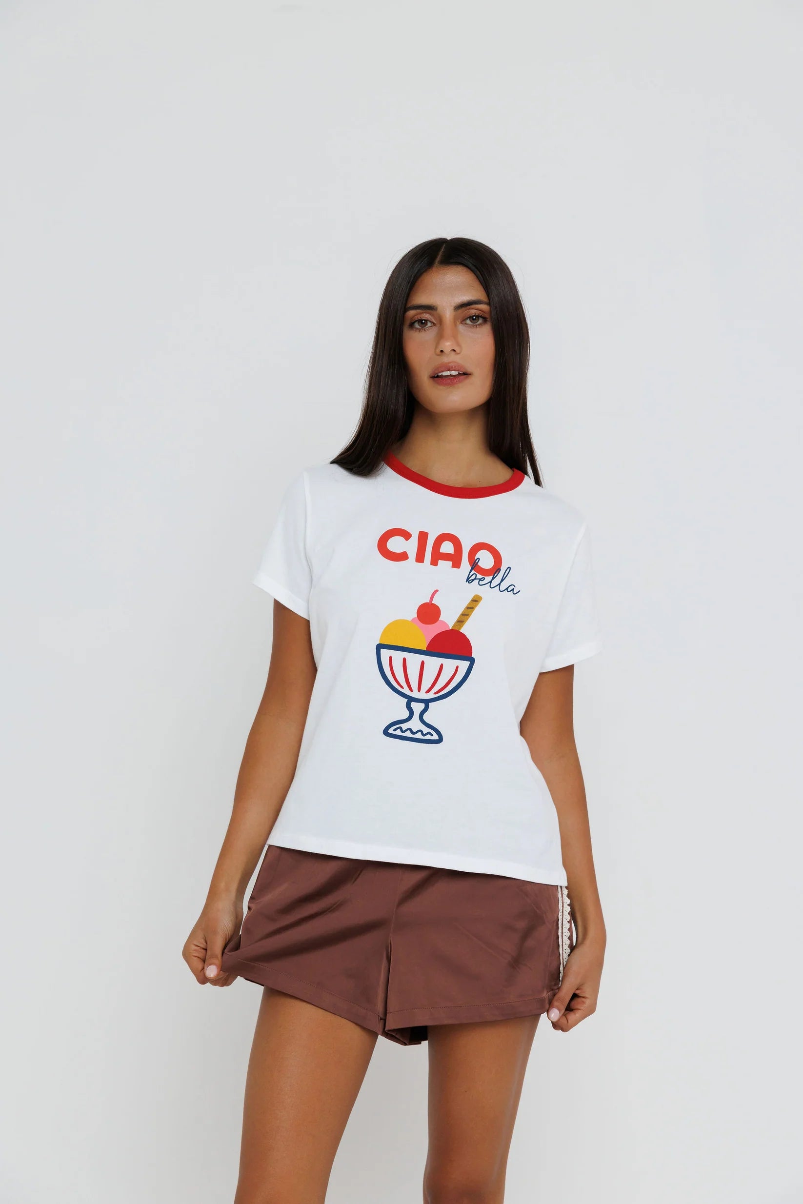 A woman modeling a white short-sleeve t-shirt with a red crew neck, featuring a colorful 'CIAO bella' ice cream sundae graphic on the front, paired with brown shorts against a light gray studio background.