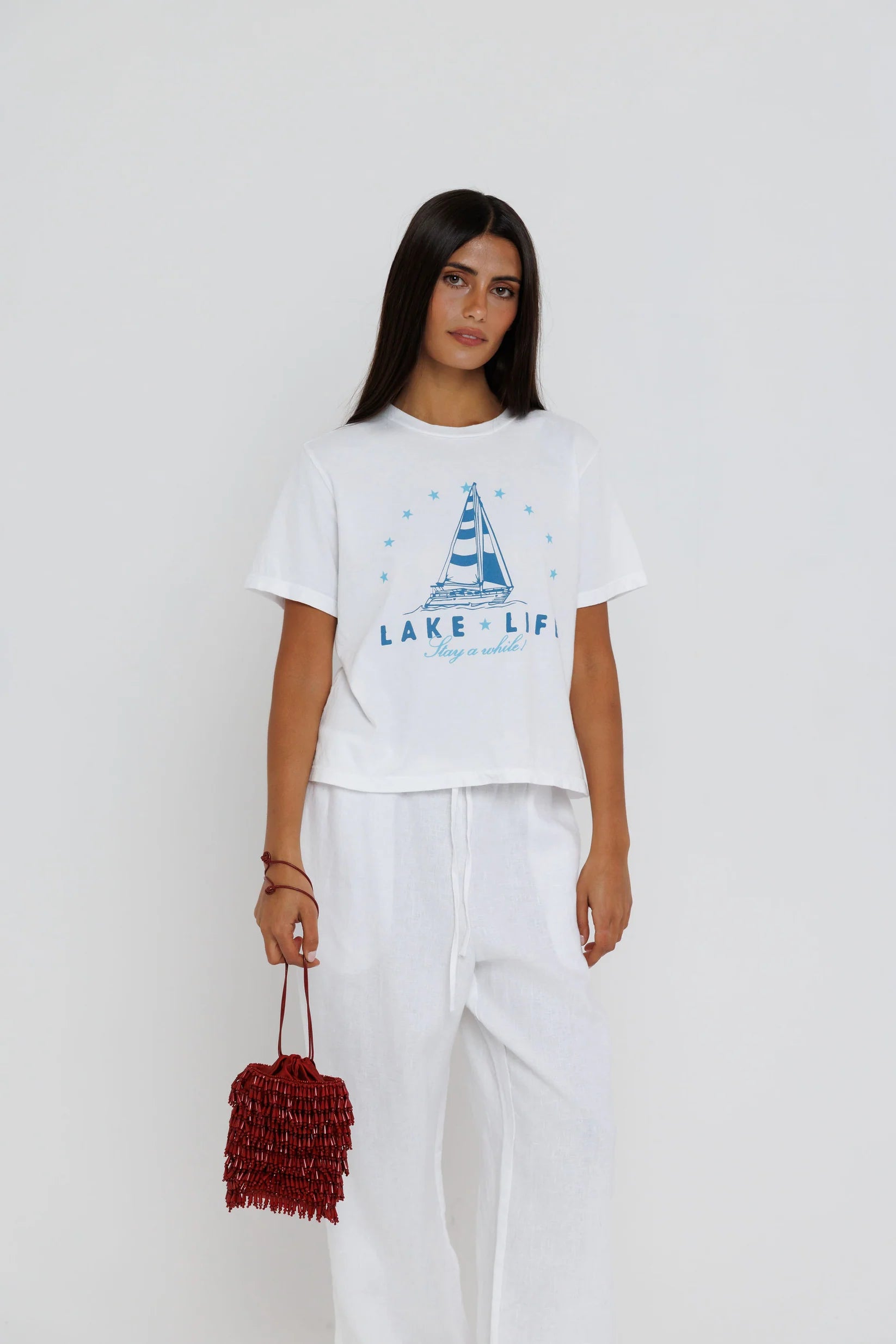 Woman wearing a white t-shirt with a sailboat design and text, holding a red bag on a white background