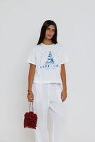 Woman wearing a white t-shirt with a sailboat design and text, holding a red bag on a white background
