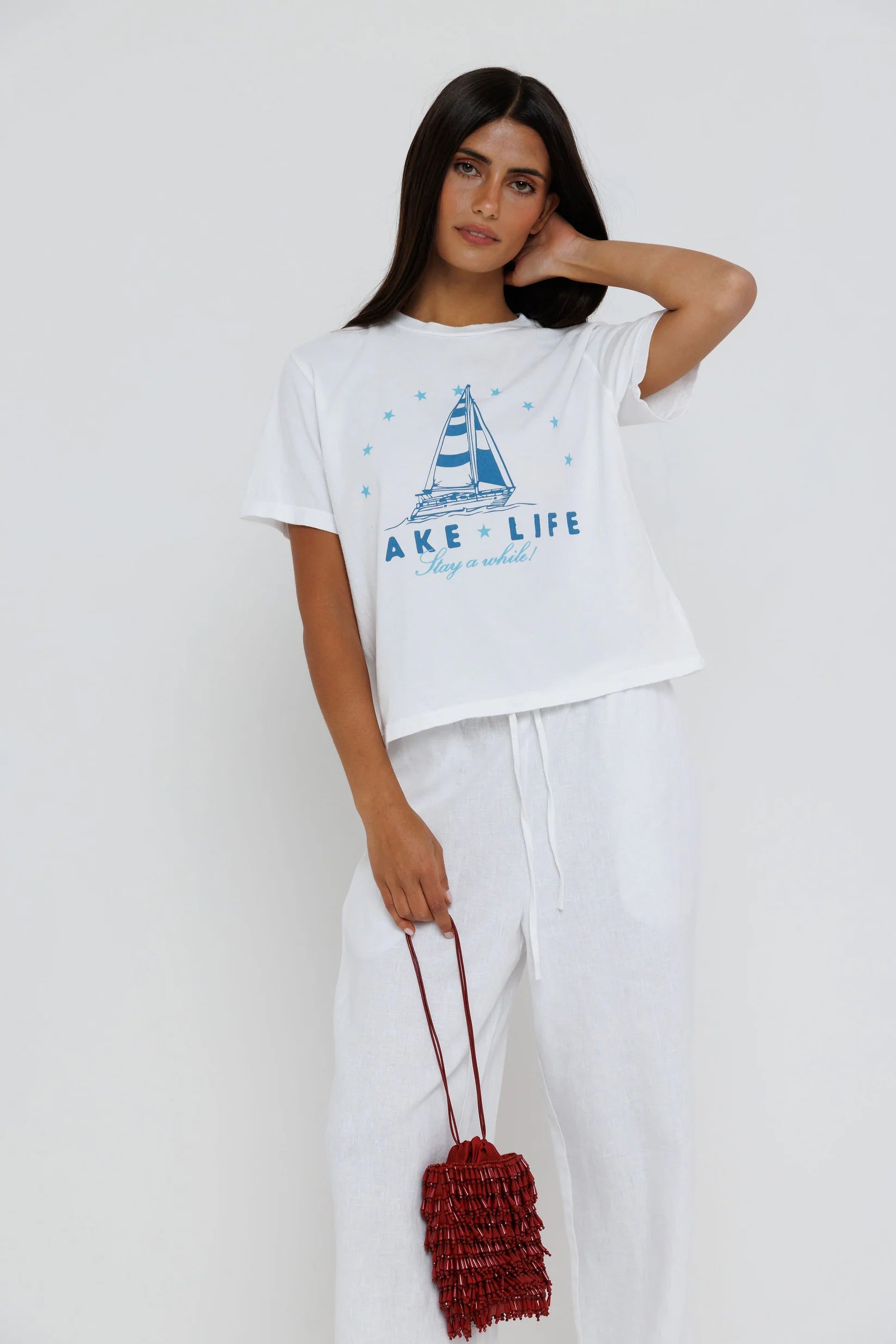 Woman wearing a white t-shirt with a sailboat graphic and 'Lake Life' text, holding a red bag against a white background.