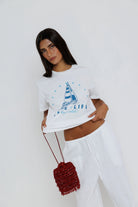 Woman wearing a white t-shirt with a sailboat design and text, holding a red bag against a white background