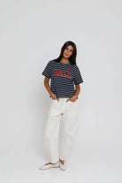 Woman wearing a striped t-shirt with a logo and white pants on a plain background