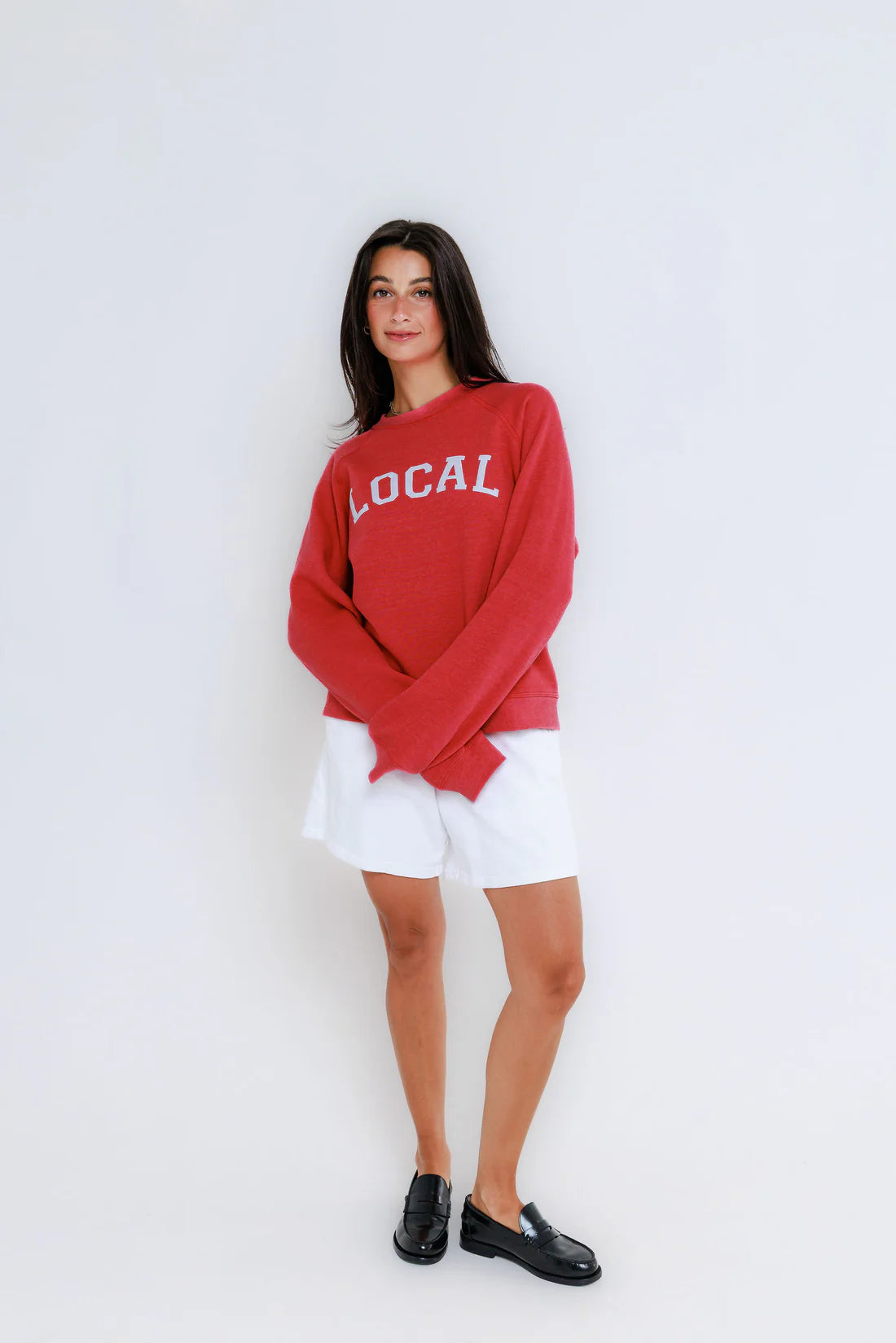 Woman wearing a red 'LOCAL' sweatshirt with white shorts on a light gray background