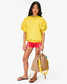 Woman wearing a yellow sweatshirt and red shorts, holding a beige bag with a yellow handle.