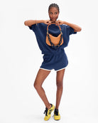 A model posing in a navy blue sweatshirt and shorts set with white trim, holding a brown woven shoulder bag, and wearing yellow sneakers, against a plain white background.