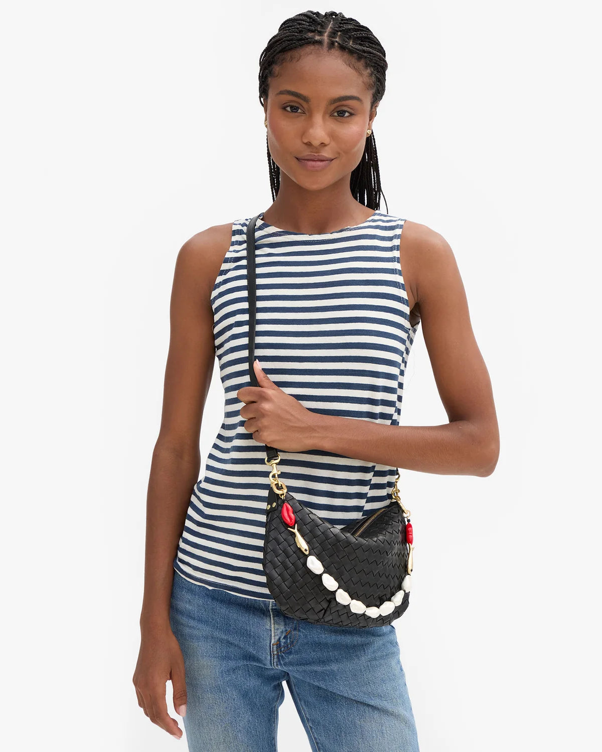 A woman wearing a navy and white striped sleeveless top and blue jeans, carrying a black woven crossbody bag with a white beaded strap and red accents against a light gray background.
