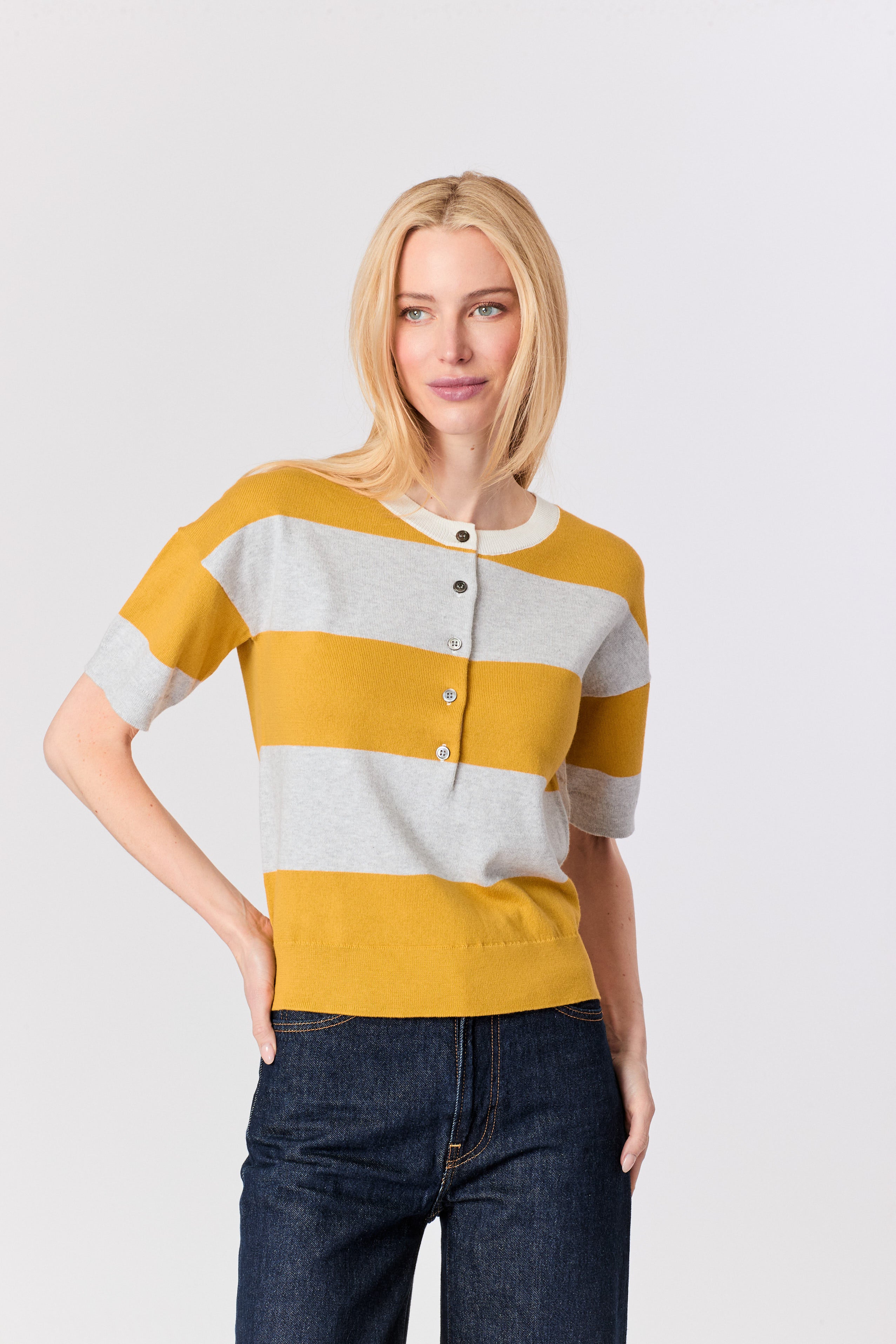A blonde woman modeling a short-sleeve knit top with horizontal yellow and light gray stripes, featuring a buttoned henley neckline, paired with dark blue jeans against a light gray studio background.
