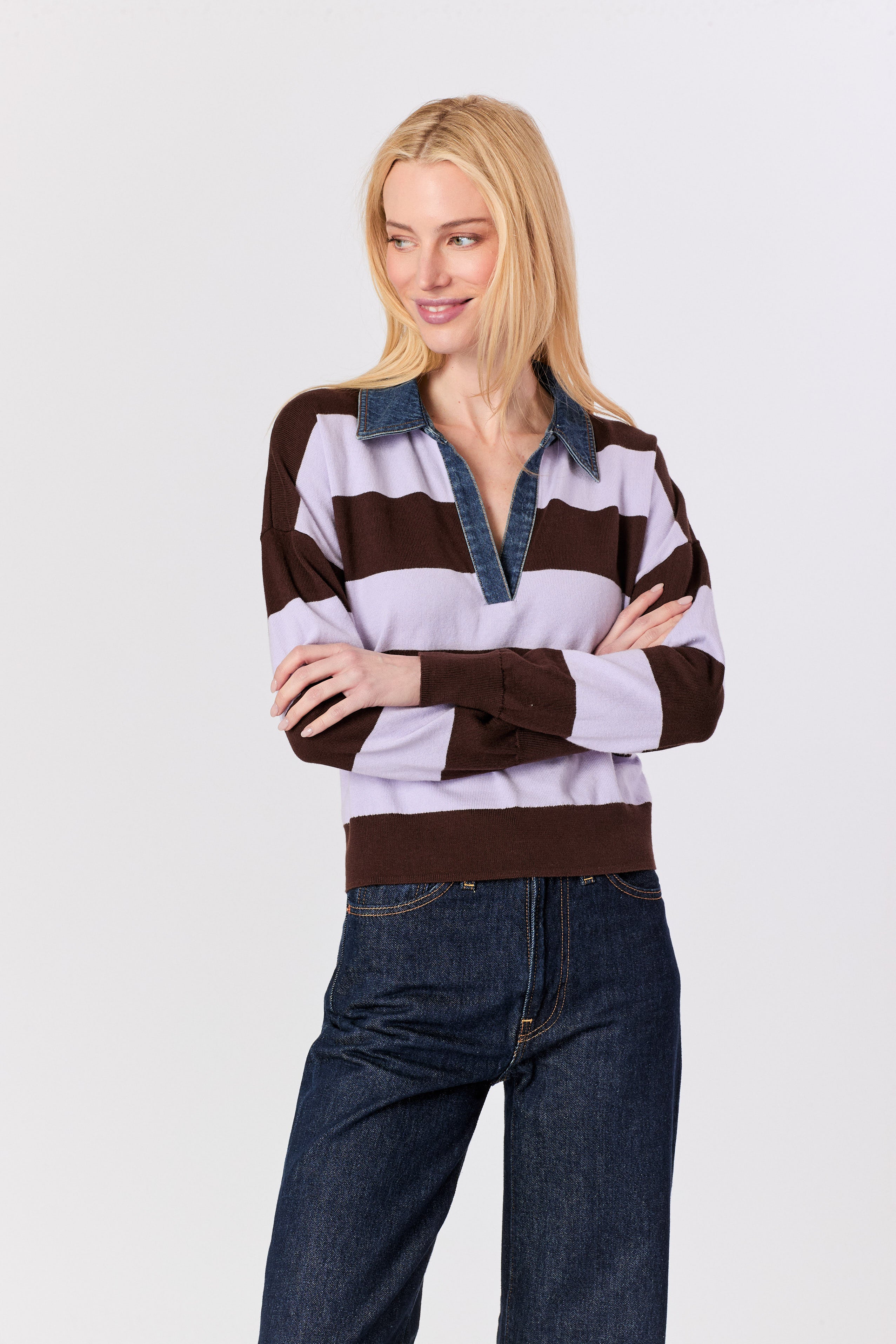 A blonde woman wearing a brown, light purple, and dark blue striped long-sleeve rugby-style sweater with dark blue jeans, standing against a light gray studio background.