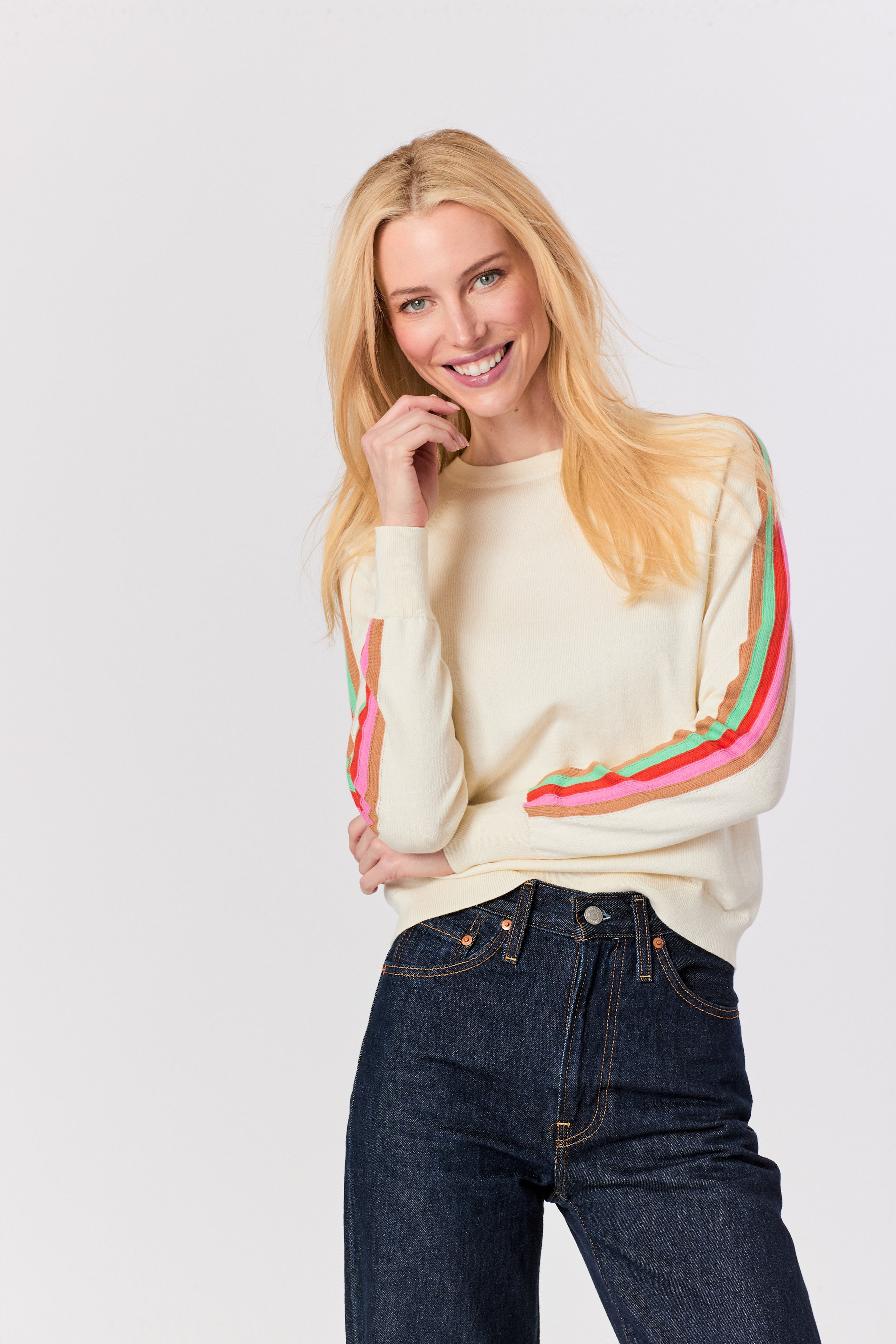 A smiling blonde woman wearing a cream-colored crewneck sweater with colorful striped details on the sleeves and dark blue jeans, photographed against a solid light gray background.