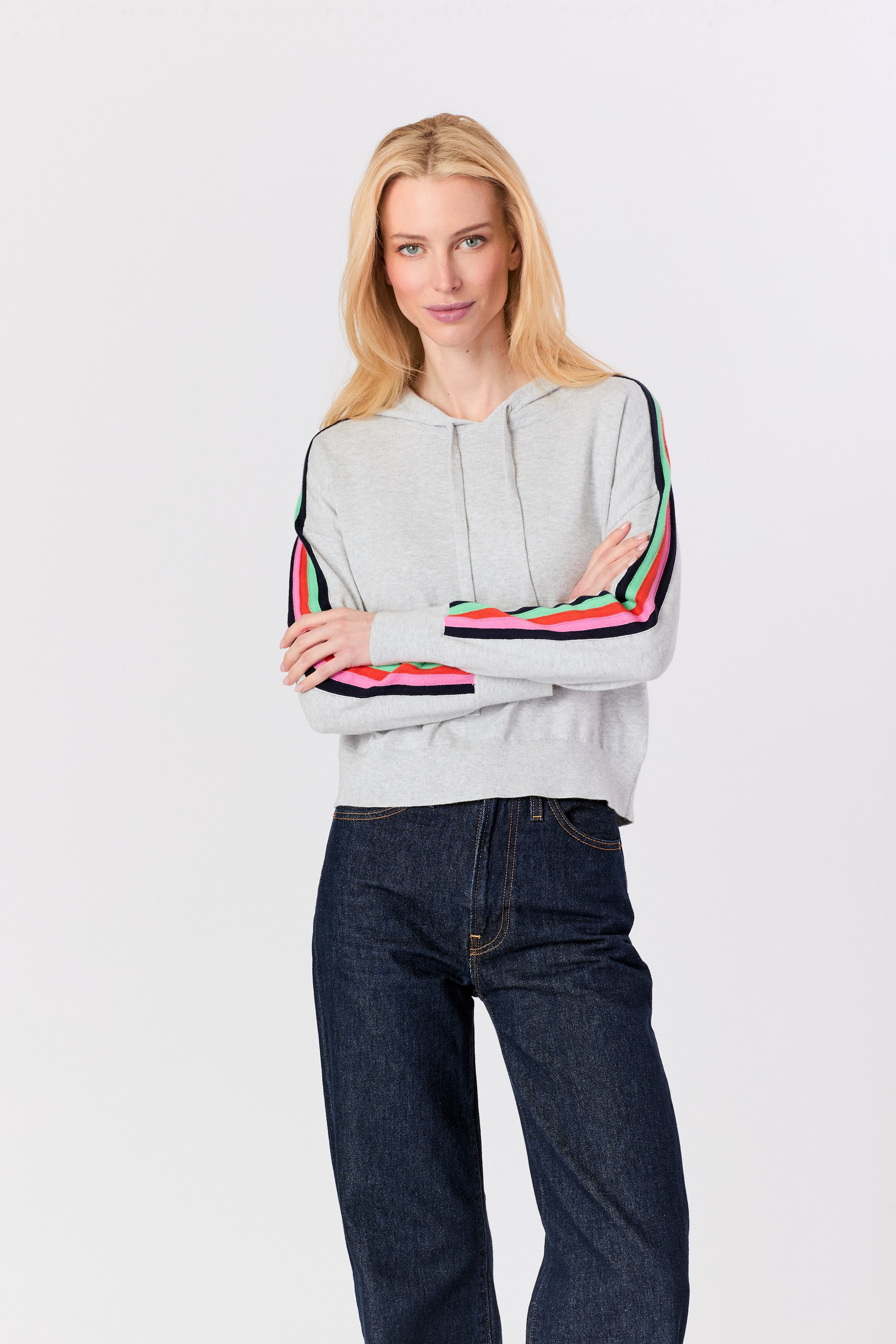 A blonde woman modeling a light gray cropped hoodie with colorful striped sleeve details, paired with dark blue jeans, against a plain light gray background.