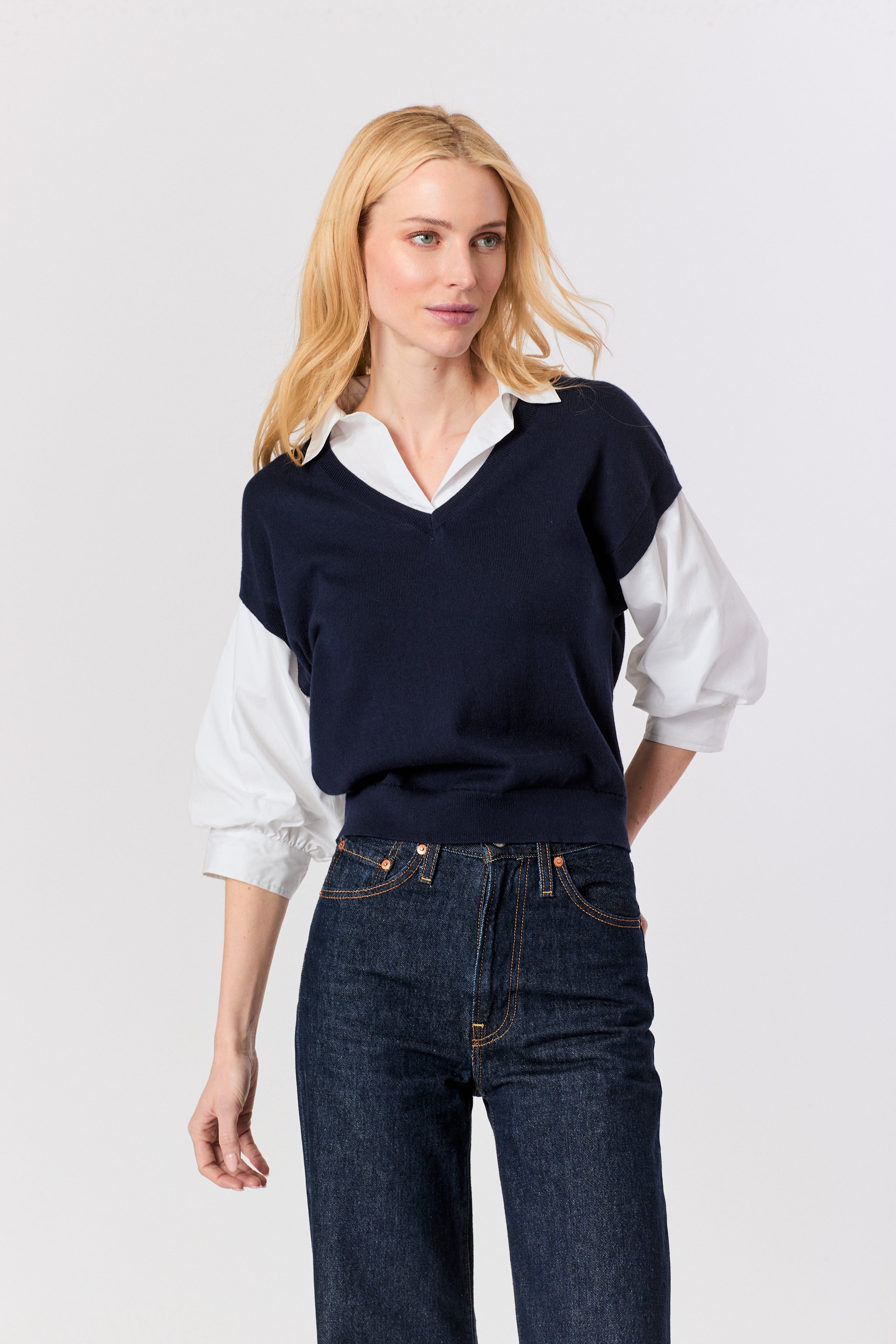 A blonde woman modeling a navy blue v-neck knit sweater vest layered over a white collared shirt and paired with dark blue jeans, against a light gray studio background.