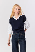 A blonde woman modeling a navy blue v-neck knit sweater vest layered over a white collared shirt and paired with dark blue jeans, against a light gray studio background.