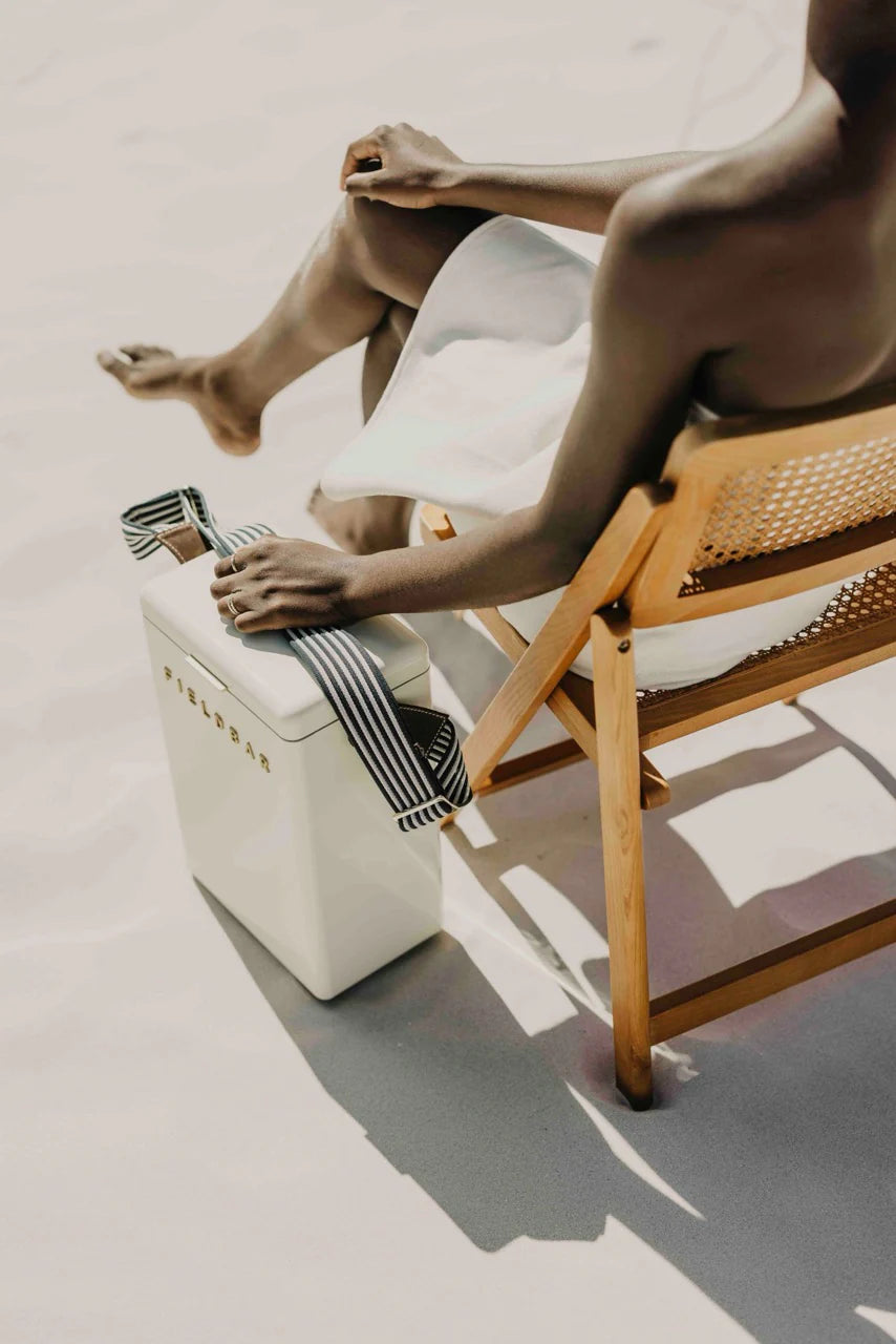 Person sitting on a wooden chair with a white ottoman and striped bag in a minimalistic setting.