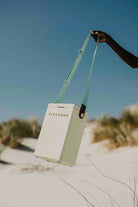 Person holding a beige cooler bag with green strap on a sandy beach.