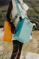 Person holding a turquoise bag with 'Fjällräven' branding in a natural setting.