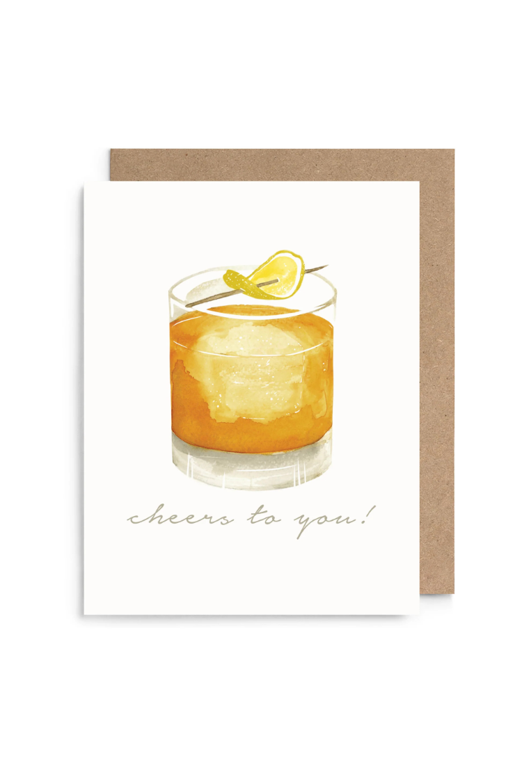 A greeting card with a watercolor illustration of an orange cocktail in a glass with a lemon garnish, above the script text 'cheers to you!'. The card is shown with a brown kraft envelope behind it on a white background.