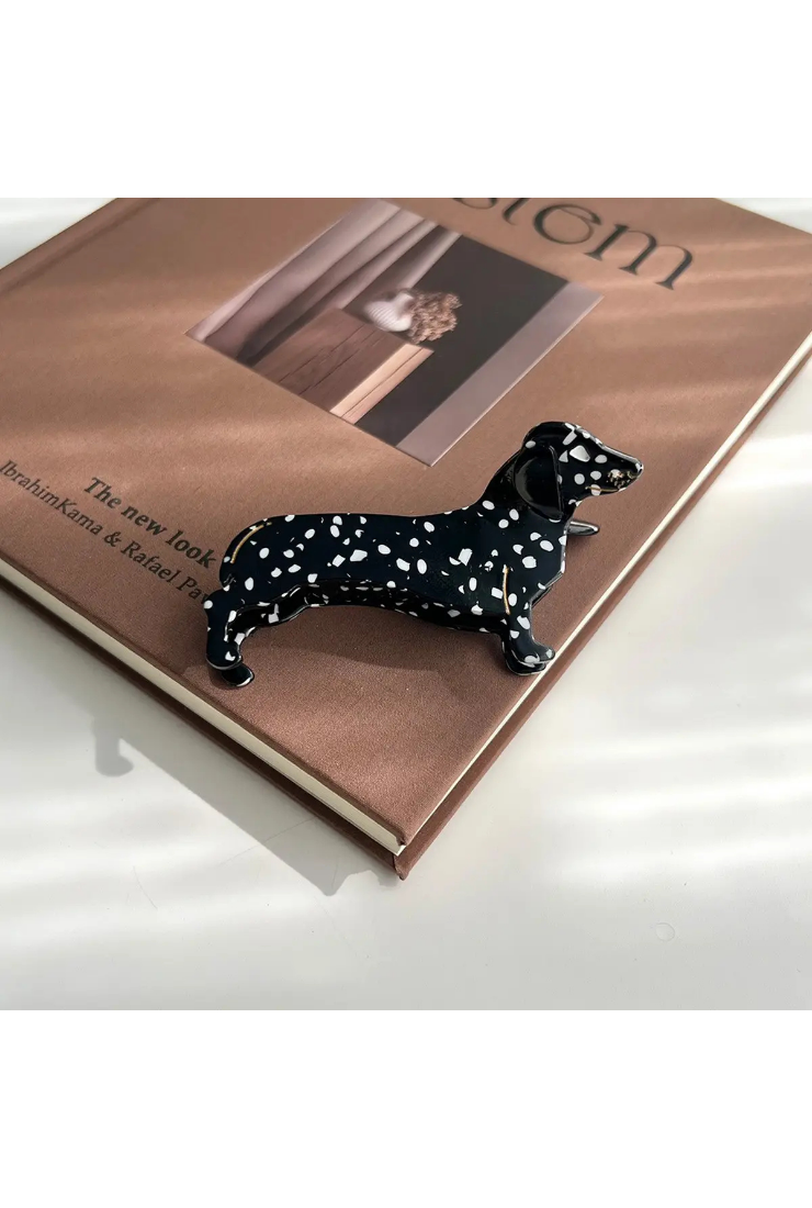 A glossy black dachshund-shaped hair claw clip with white spots, resting on the edge of a brown book on a white surface.