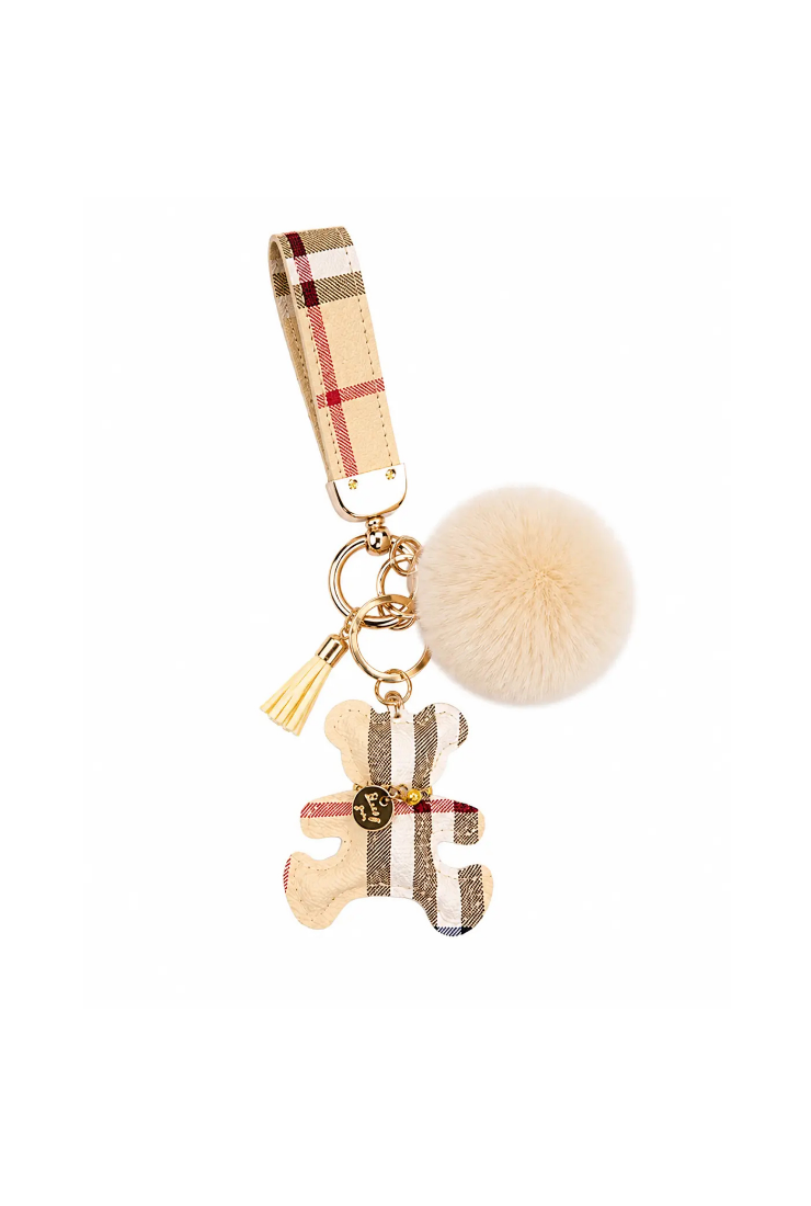 A beige and brown plaid keychain charm featuring a puzzle piece charm, a fluffy cream pom-pom, a small gold tassel, and a gold-tone ring with a small gold logo medallion.