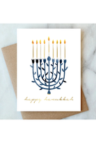 A white Hanukkah greeting card featuring a navy blue menorah with lit gold candles and the words 'happy hanukkah' in gold script, resting on a brown kraft envelope over a light marble surface.