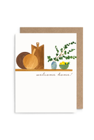 A white greeting card with a watercolor-style illustration of wooden cutting boards, a bowl, greenery, and a small yellow bowl, with the words 'welcome home!' printed below. A kraft brown envelope is placed behind the card on a white background.