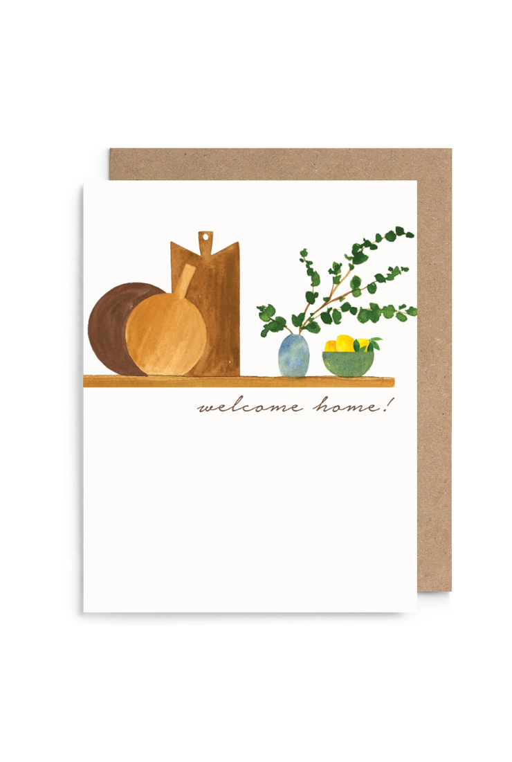A white greeting card with a watercolor-style illustration of wooden cutting boards, a bowl, greenery, and a small yellow bowl, with the words 'welcome home!' printed below. A kraft brown envelope is placed behind the card on a white background.