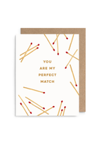 A white greeting card with gold text reading 'YOU ARE MY PERFECT MATCH' and scattered gold matchstick illustrations with red tips, shown with a kraft brown envelope on a white background.