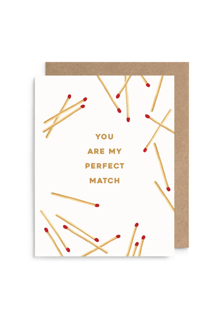 A white greeting card with gold text reading 'YOU ARE MY PERFECT MATCH' and scattered gold matchstick illustrations with red tips, shown with a kraft brown envelope on a white background.