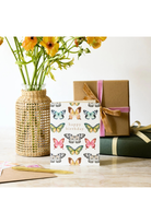 A white 'Happy Birthday' greeting card decorated with colorful butterfly illustrations, styled on a tabletop next to a brown gift box with a pink ribbon, a green wrapped present, and a wicker vase with yellow flowers.