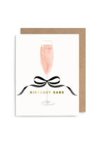 A square birthday card with a kraft envelope, featuring an illustration of a champagne flute filled with pink bubbly drink and a black ribbon bow, with the text 'BIRTHDAY BABE' in gold.