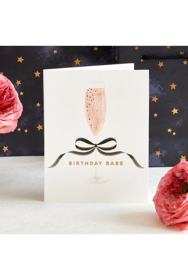 A white greeting card with a champagne flute illustration, pink bubbly liquid, a black ribbon bow, and the text 'BIRTHDAY BABE' in gold, styled with pink roses against a dark starry background.