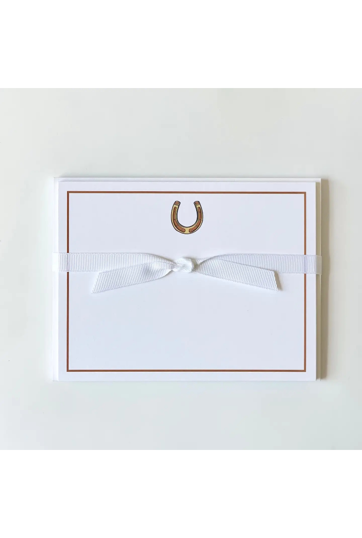 A white square greeting card or note card with a thin brown border, a small gold horseshoe motif near the top center, and a white ribbon tied in a bow across the middle, shown on a light neutral background.