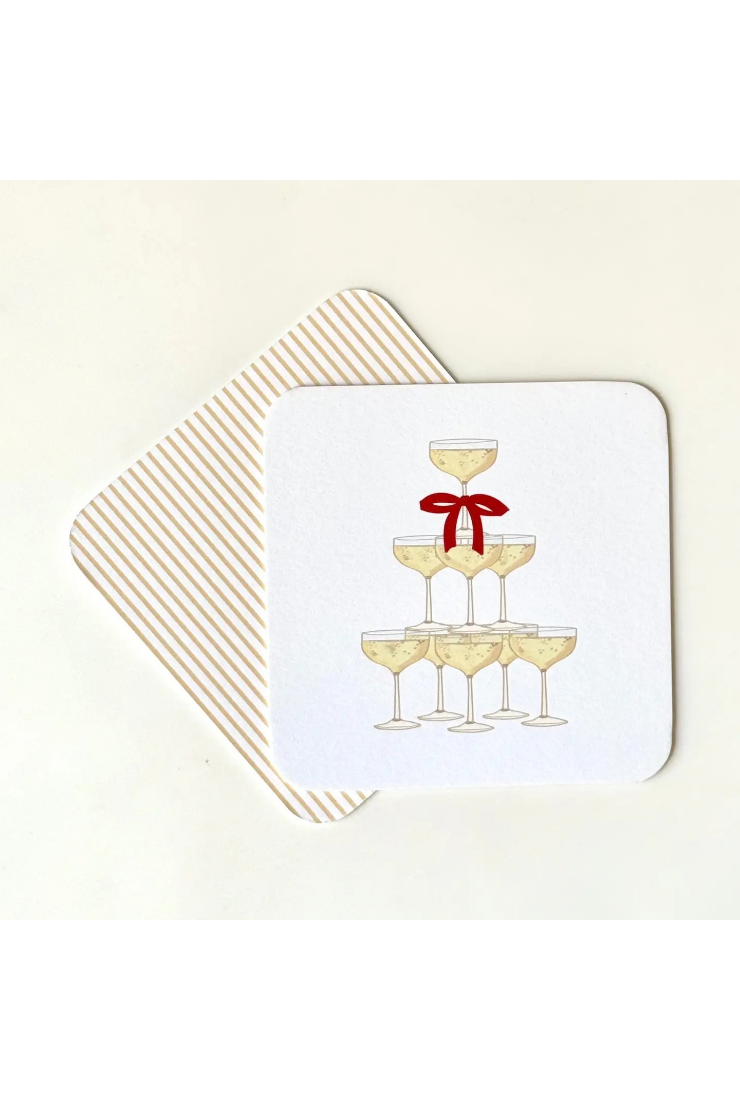 A square white coaster with rounded corners featuring an illustration of a champagne tower with a red bow, placed on top of a striped square coaster against a light cream background.