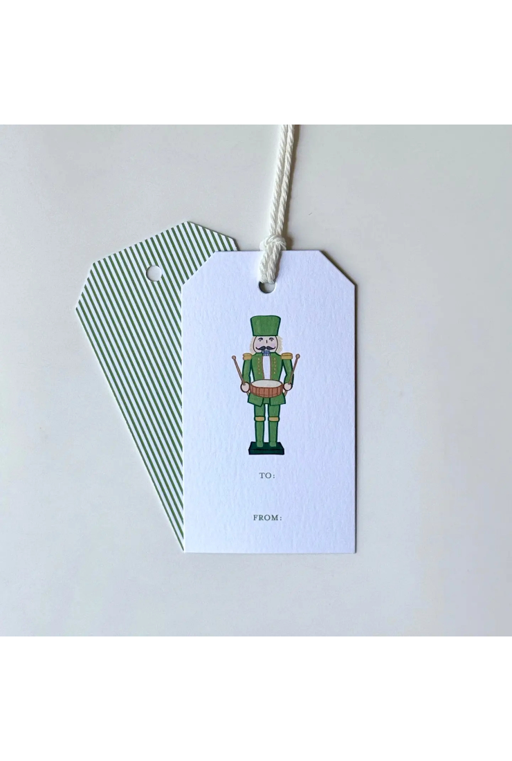 A white gift tag with a printed nutcracker illustration and the words 'TO:' and 'FROM:' hangs from a white string, with a second green-and-white striped tag partially visible behind it on a light gray background.