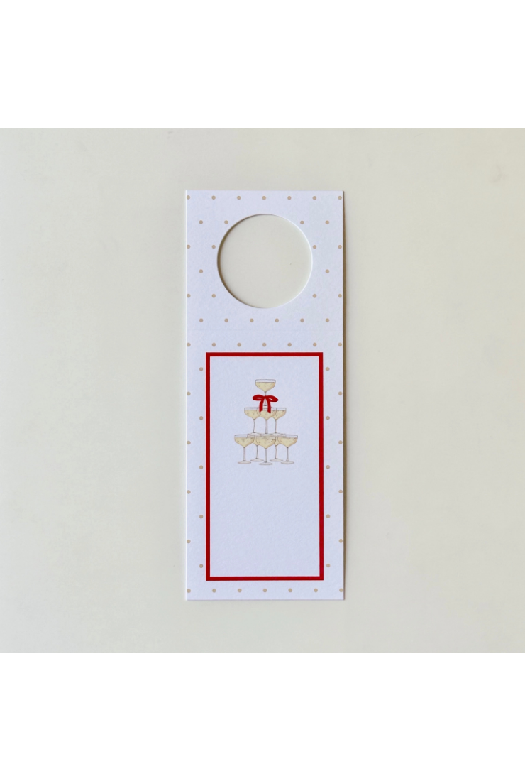 A white hanging hotel doormat with a circular cutout at the top, decorated with a red border, a red bow, champagne glasses, and small gold polka dots on a light background.