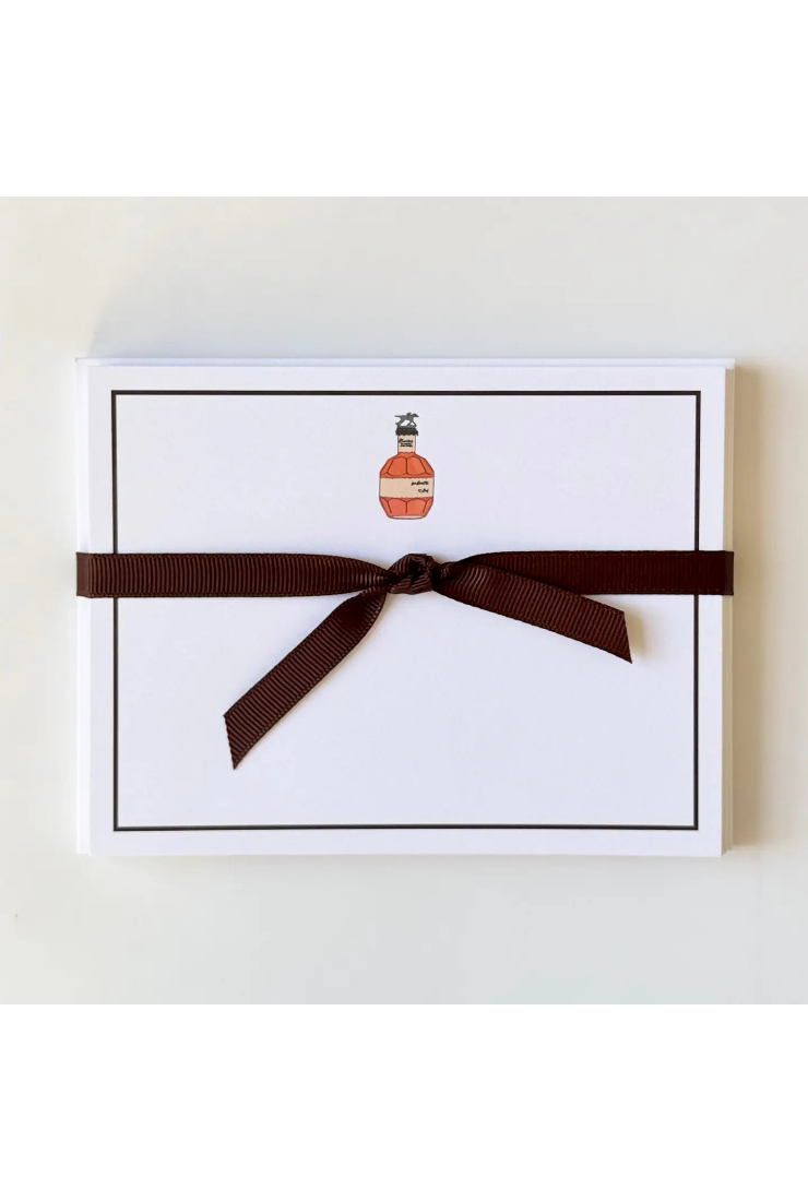 A white rectangular gift box or package tied with a dark brown ribbon, featuring a small illustrated bottle graphic near the top center, photographed against a light neutral background.