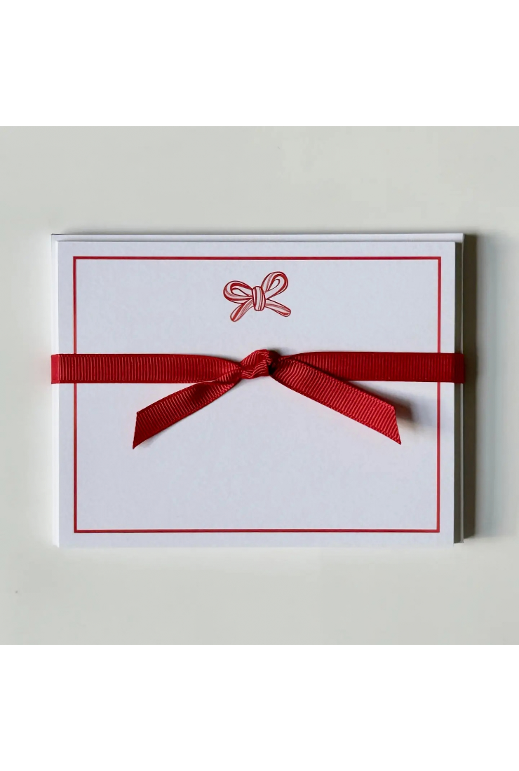 A white square greeting card or note pad with a thin red border, a small red bow illustration near the top, and a red ribbon tied in a bow across the front, shown on a light neutral background.
