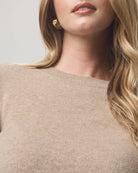 Close-up of a woman wearing a ribbed knit crewneck sweater in a light beige color, with a small gold earring visible against a neutral light gray background.