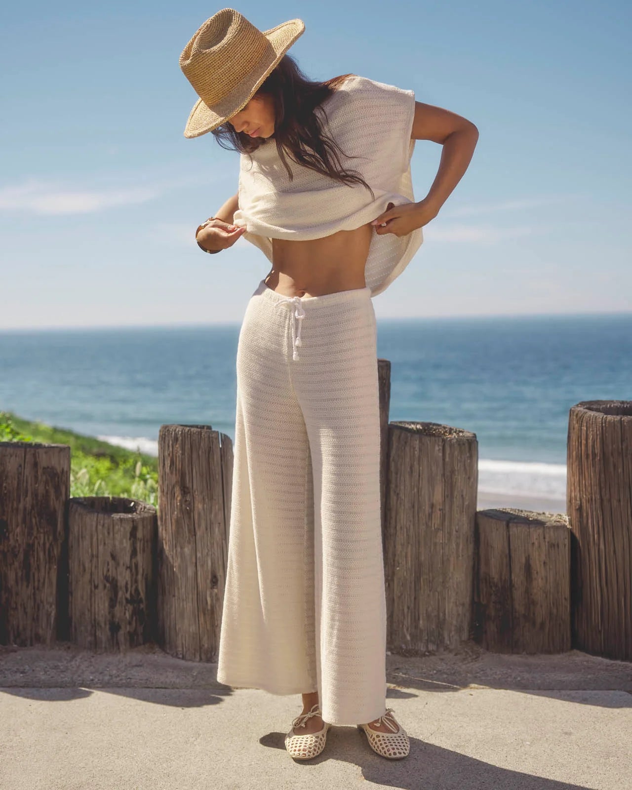 Woman in a white outfit with a straw hat standing by the ocean.