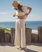Woman in a white outfit with a straw hat standing by the ocean.
