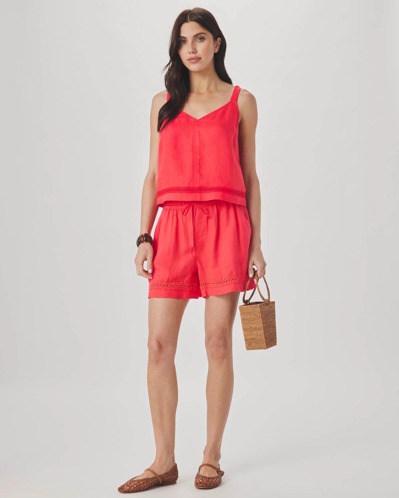 Woman wearing a coral sleeveless top and shorts set, holding a woven bag on a white background