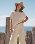 Woman in a white outfit and straw hat standing by a wooden post with ocean view