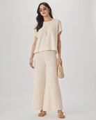 Woman wearing a beige outfit with wide-leg pants and a top, holding a straw bag on a light gray background.