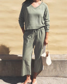 A woman wearing a matching sage green lounge set consisting of a long-sleeve V-neck top and drawstring cropped pants, holding a cream-colored tote bag against a textured beige wall.