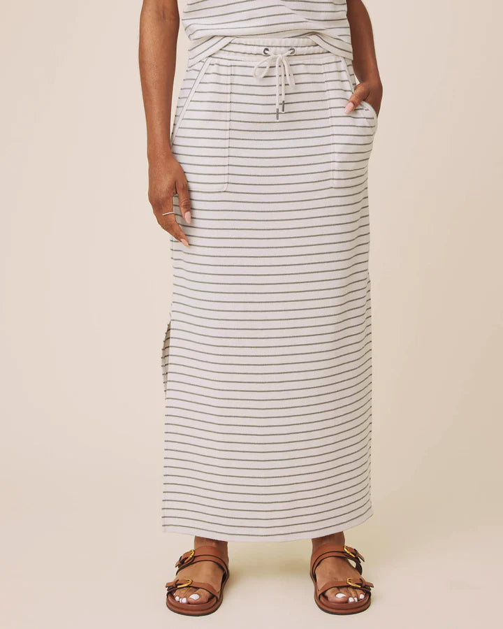 A person wearing a long white maxi skirt with thin dark horizontal stripes, side pockets, and a drawstring waist, paired with brown strappy sandals against a plain beige studio background.