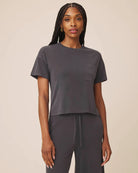 A woman modeling a dark gray short-sleeve crop top with a chest pocket, paired with matching dark gray drawstring pants, against a solid beige background.